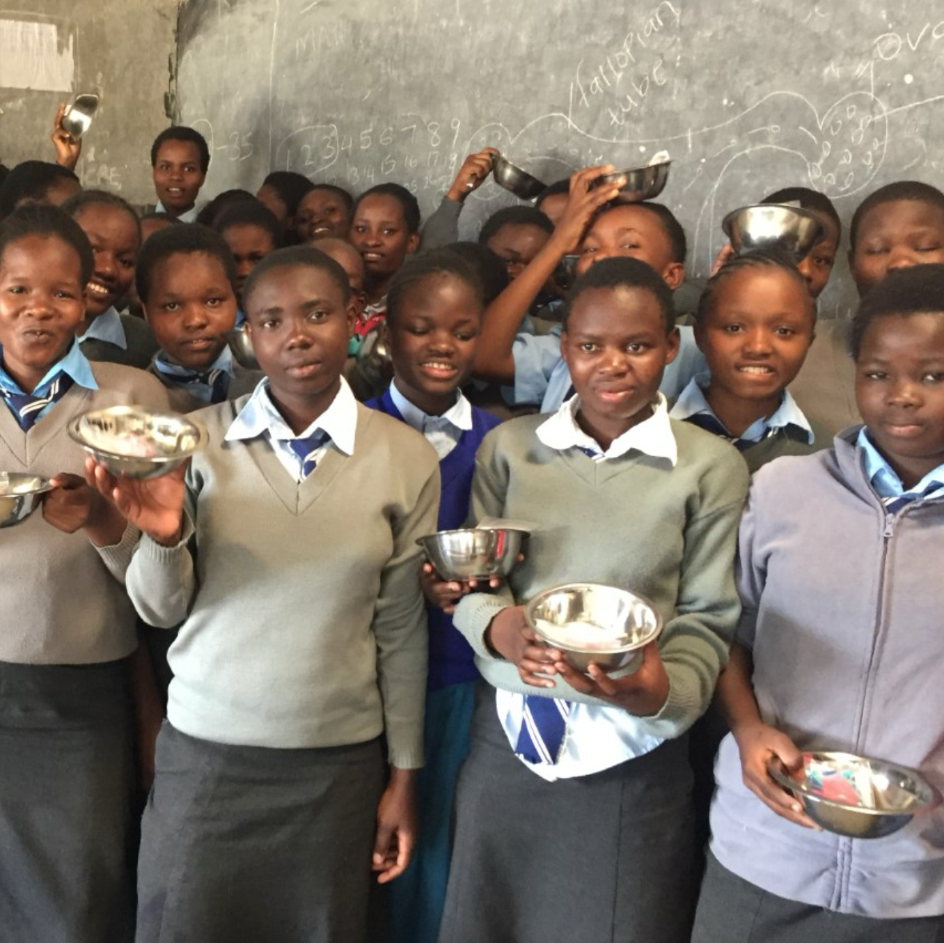 Children holding metal bowls in a classroom setting with a chalkboard in the background.