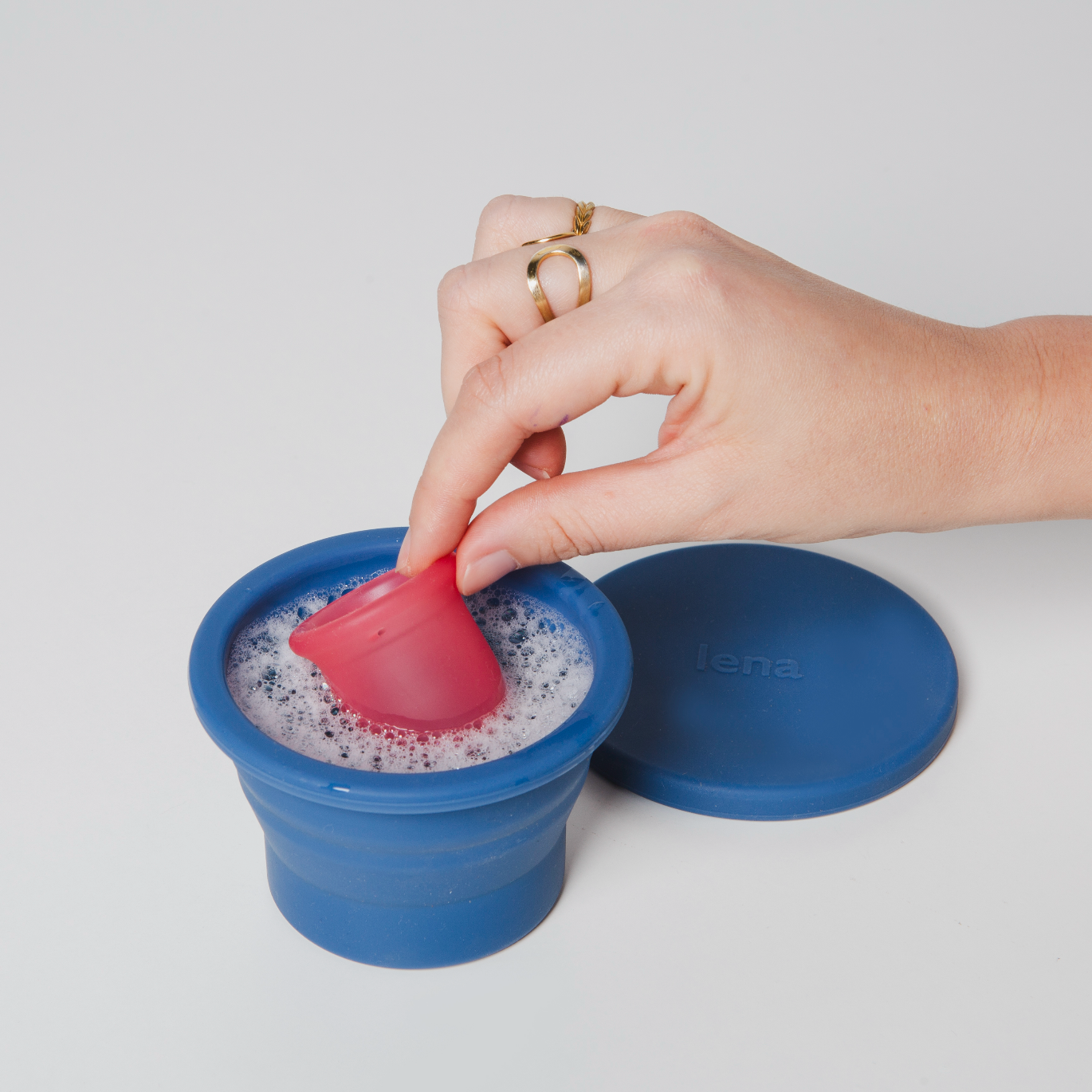 Hand using a pink silicone cup to clean a blue Lena Cleaning Container with soap suds on a light gray background.