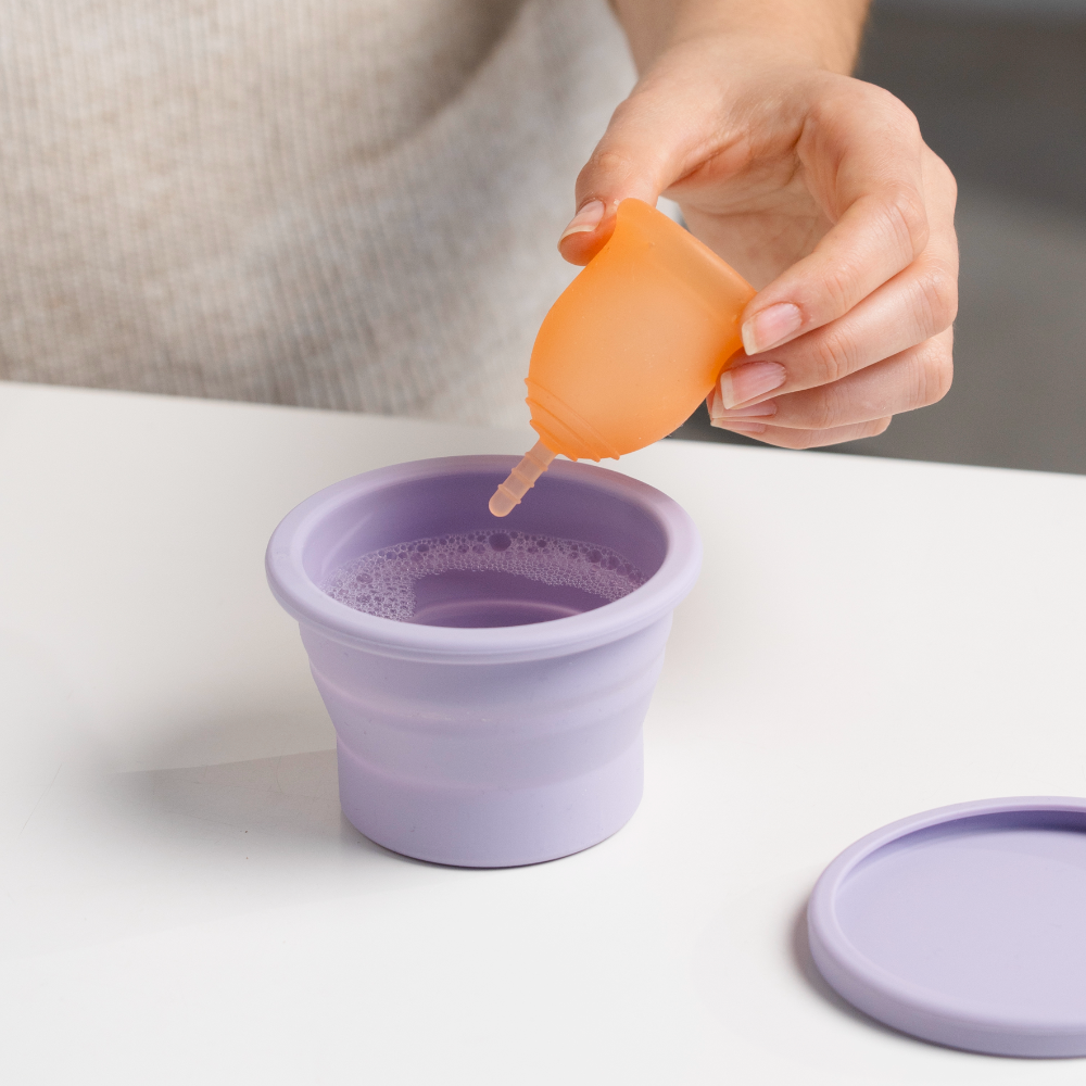 Hand holding a coral soft lena Cup over purple Lena Cup Cleaning Container to sanitize it.