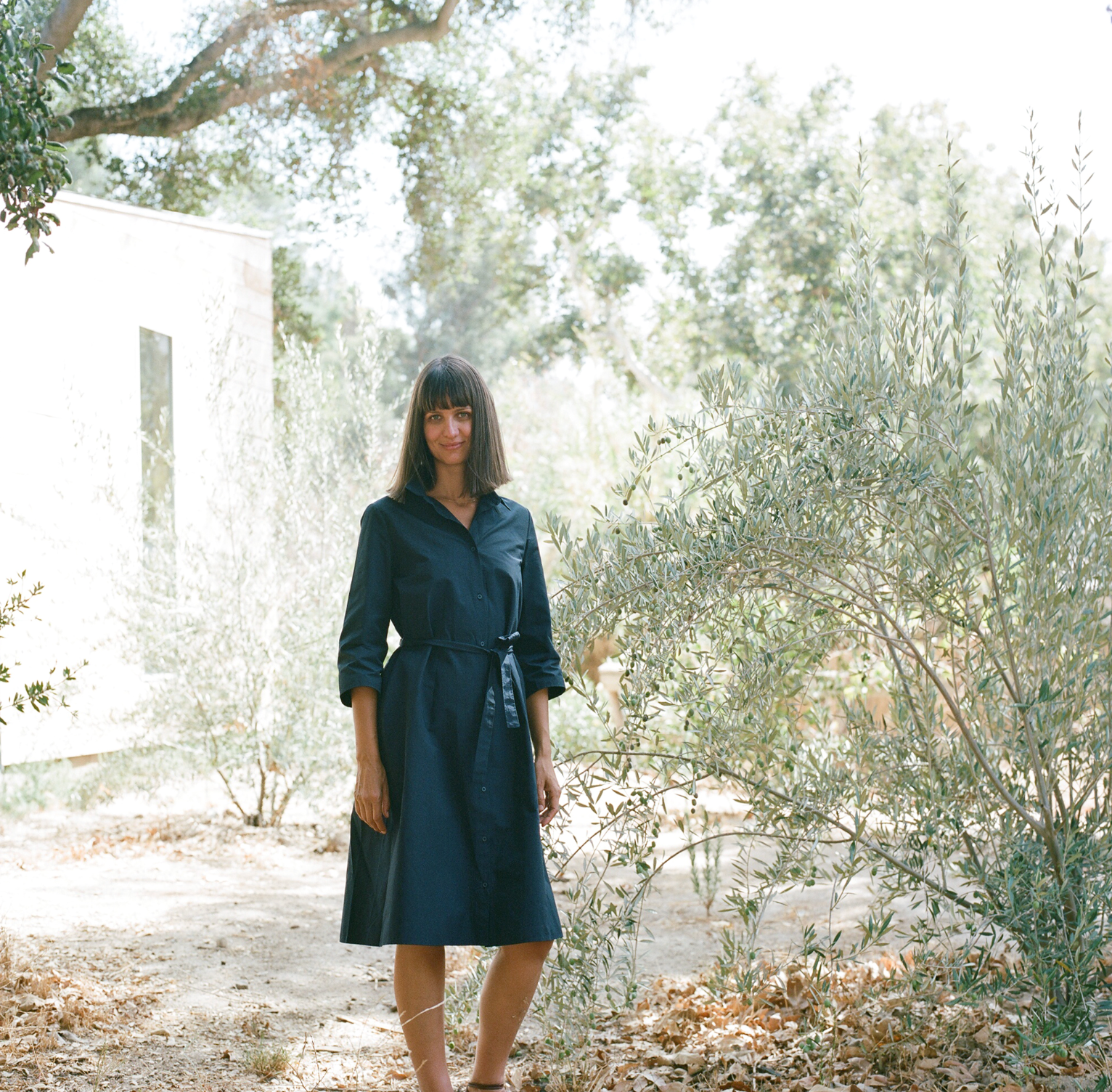 Woman wearing a navy dress stands in dry country side, next to olive tree