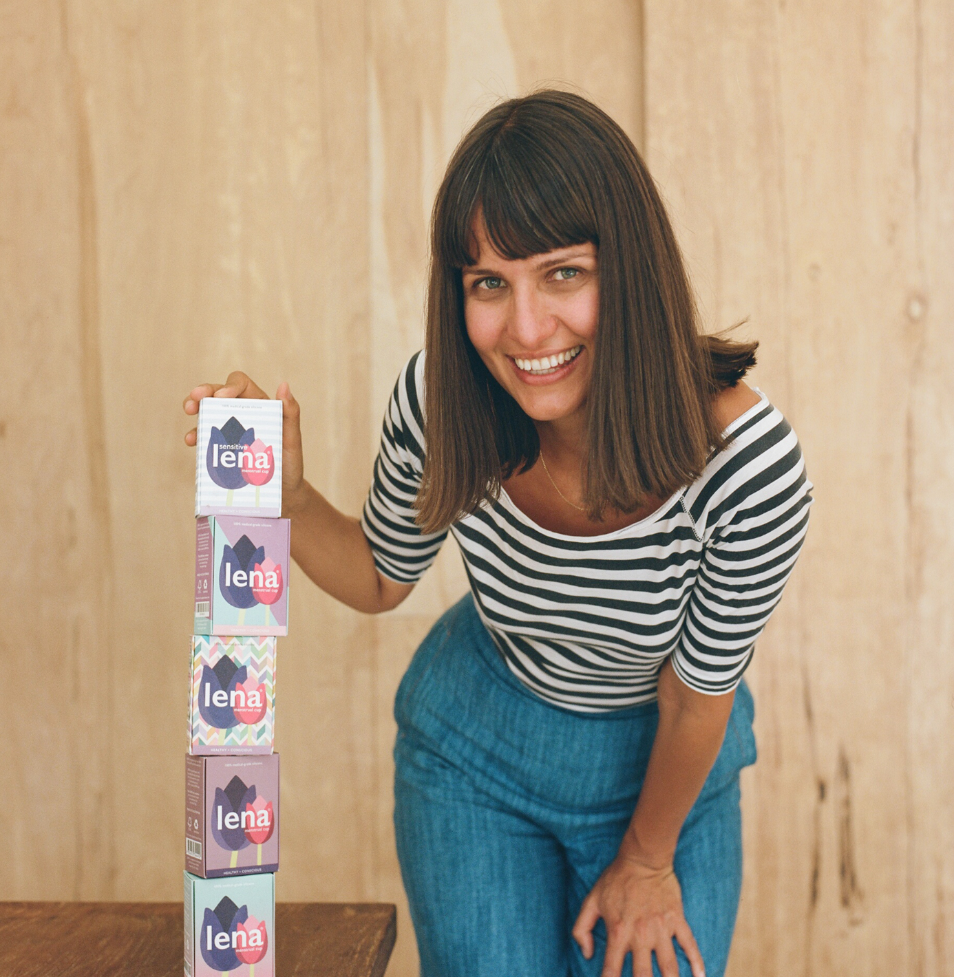 Woman with brown hair is looking at the camera, wearing black-and-white striped shirt and jeans, holding a tower of Lena Cup boxes