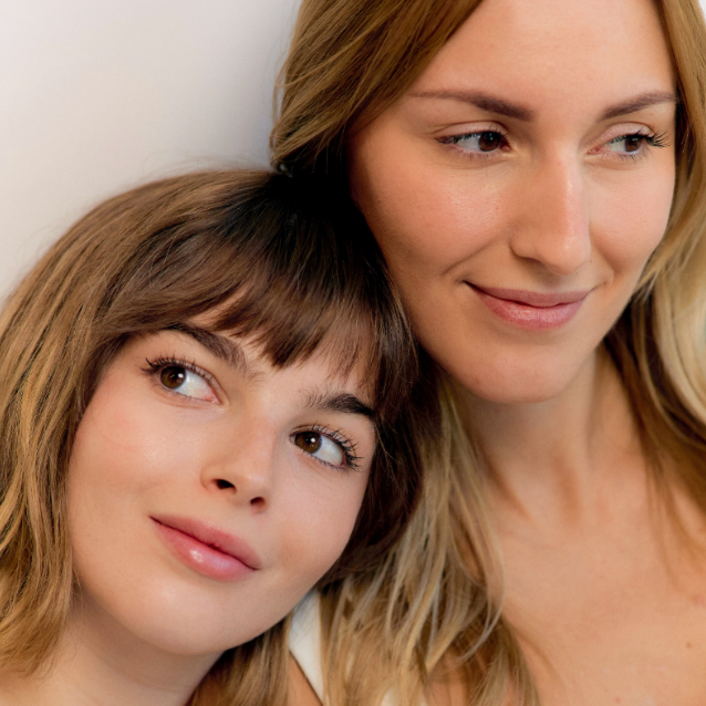 Two women with light brown hair posing closely together against a neutral background